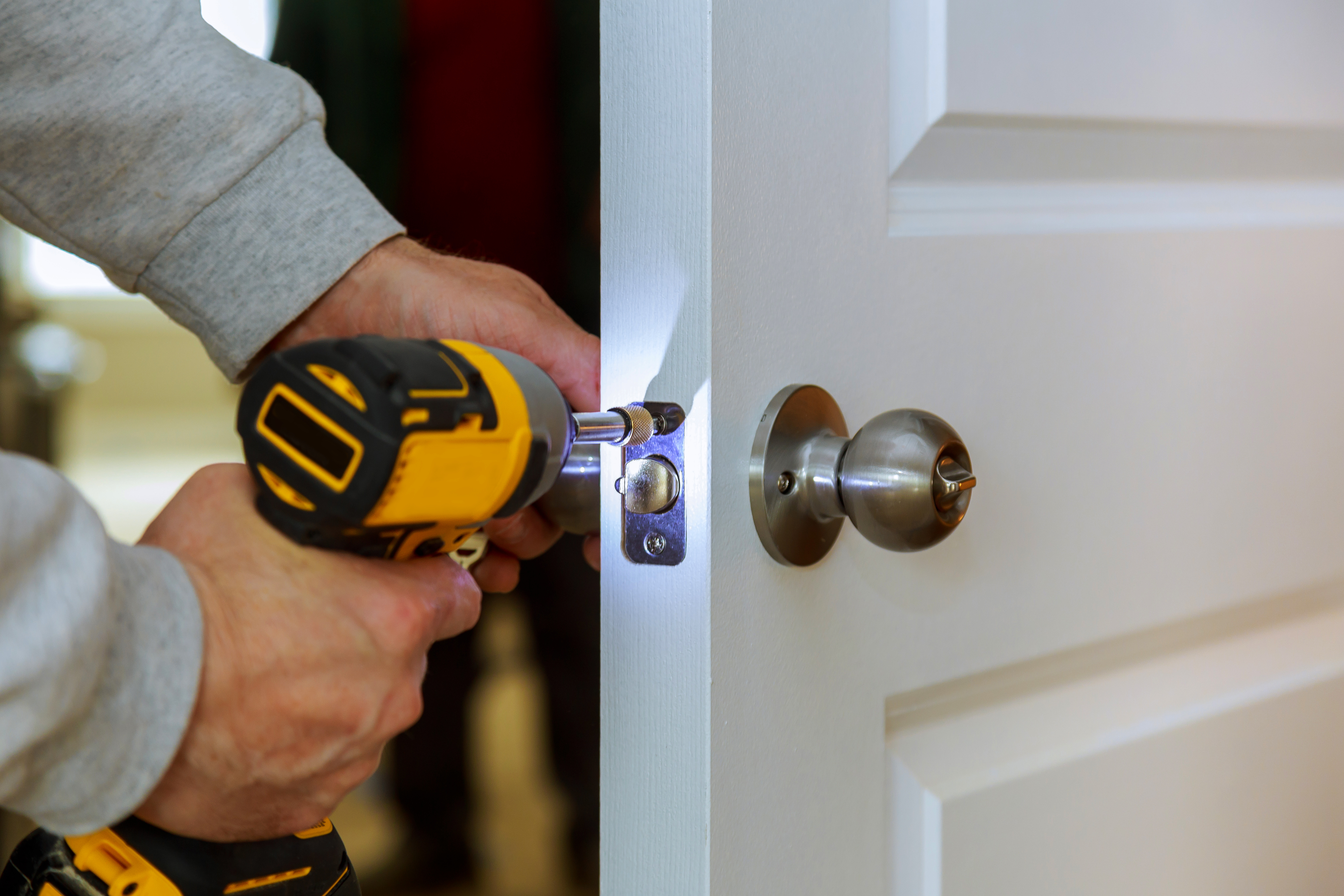 A person installing a doorknob on a white door using a cordless drill.