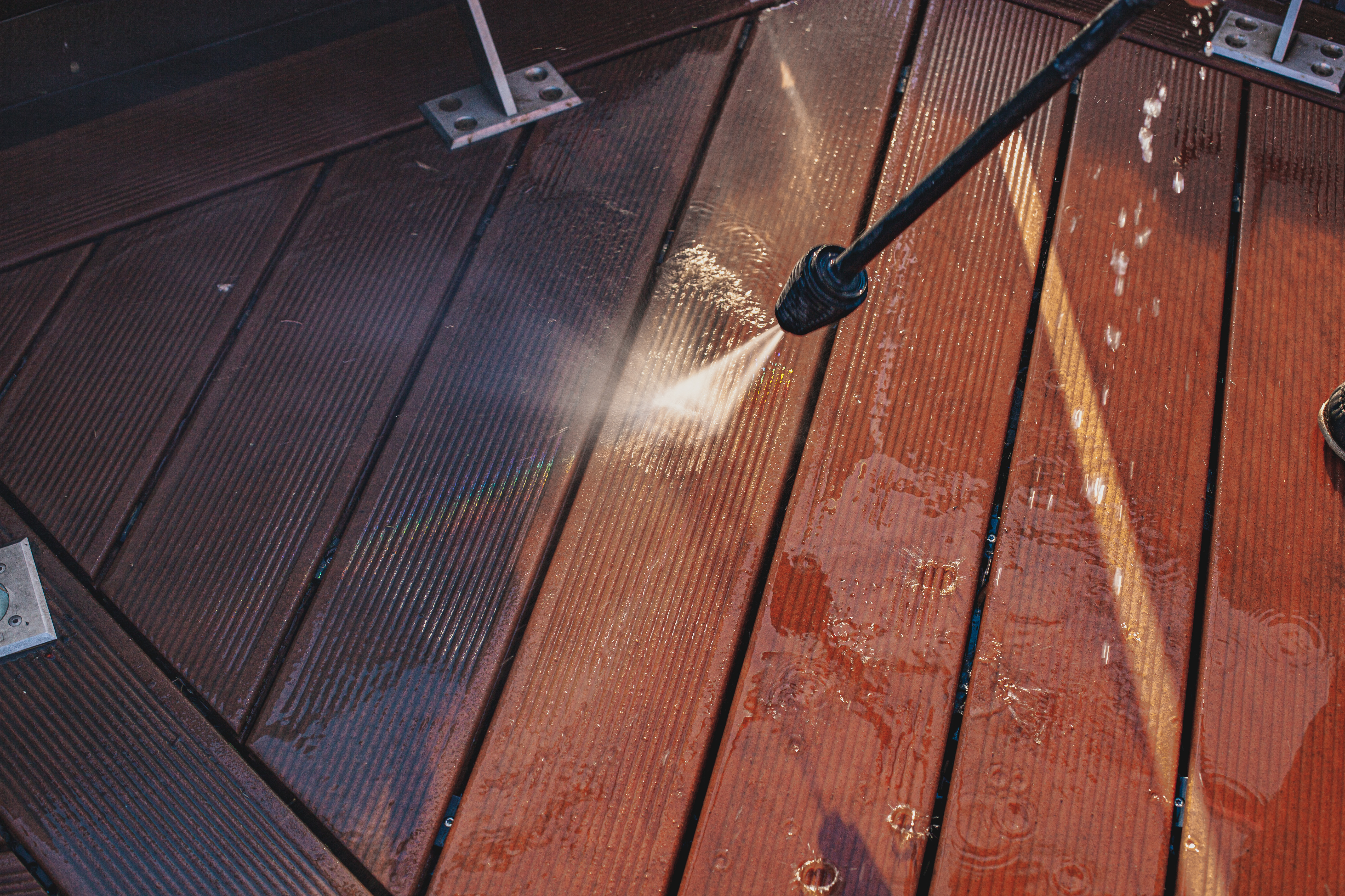 Close-up of a power washer cleaning a wooden deck, showing water spraying and wood brightening.