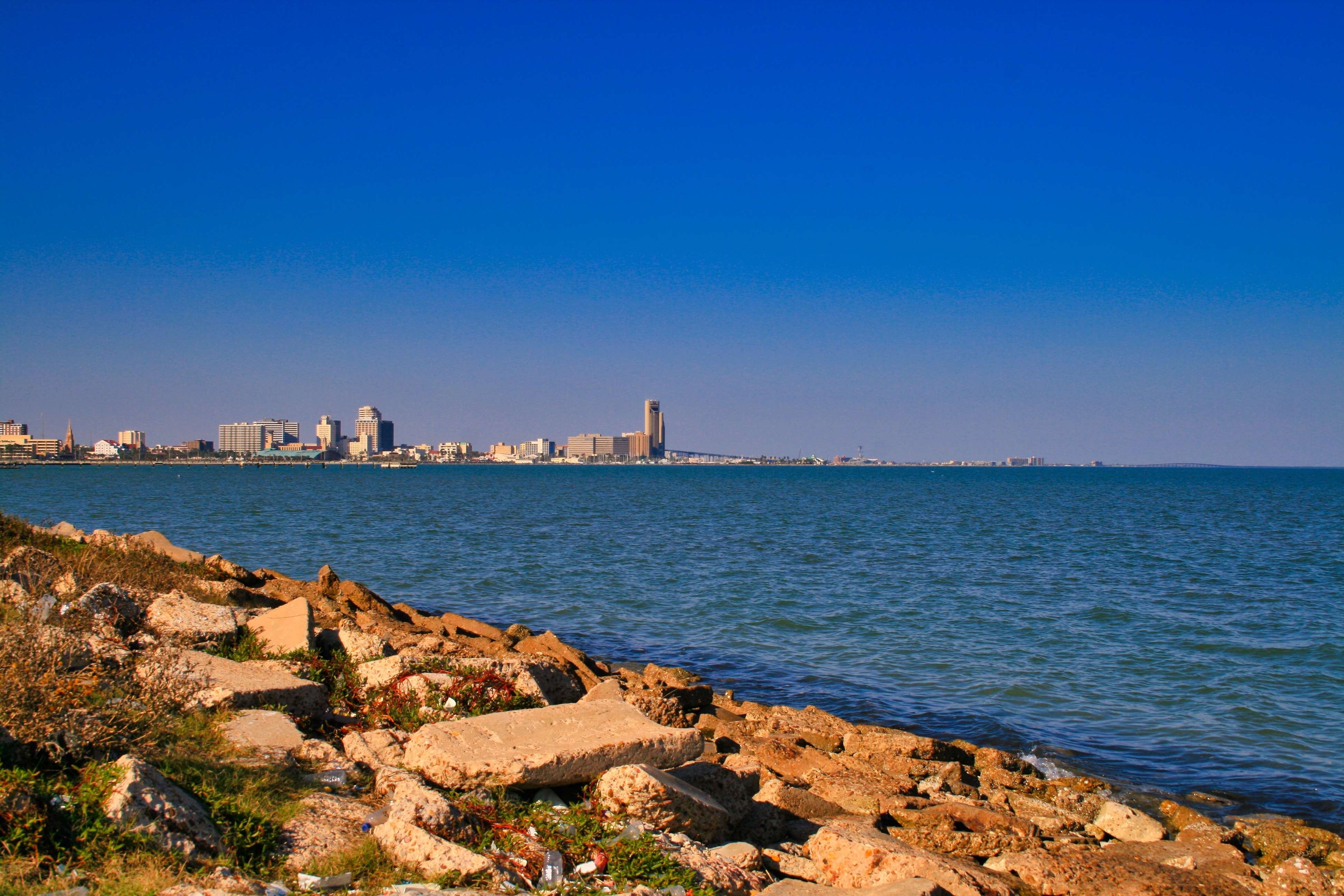 Coastal view of Corpus Christi with a clear blue sky and urban skyline from a rocky shoreline.