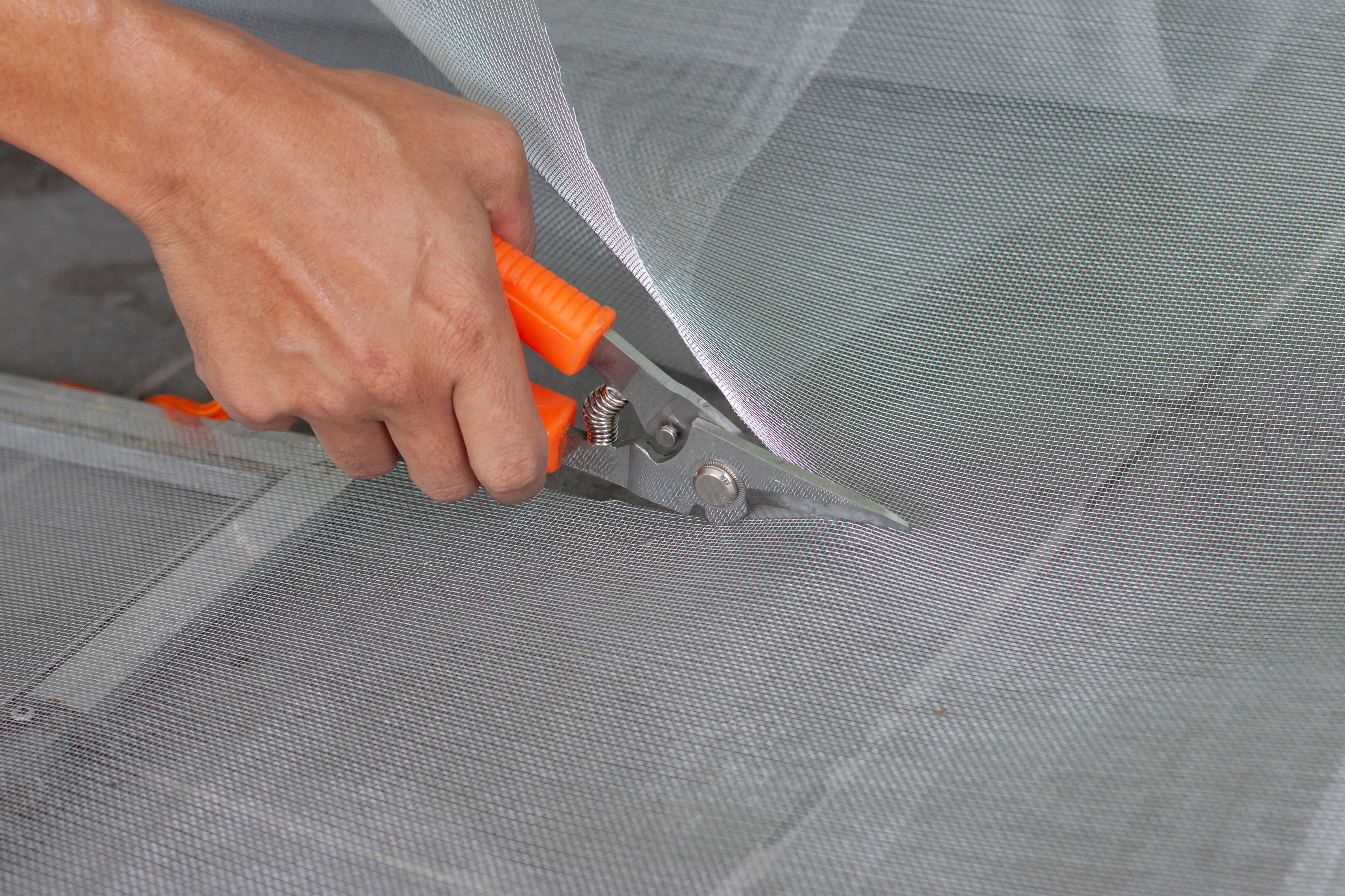 Hands cutting window screen mesh with orange scissors during repair preparation.