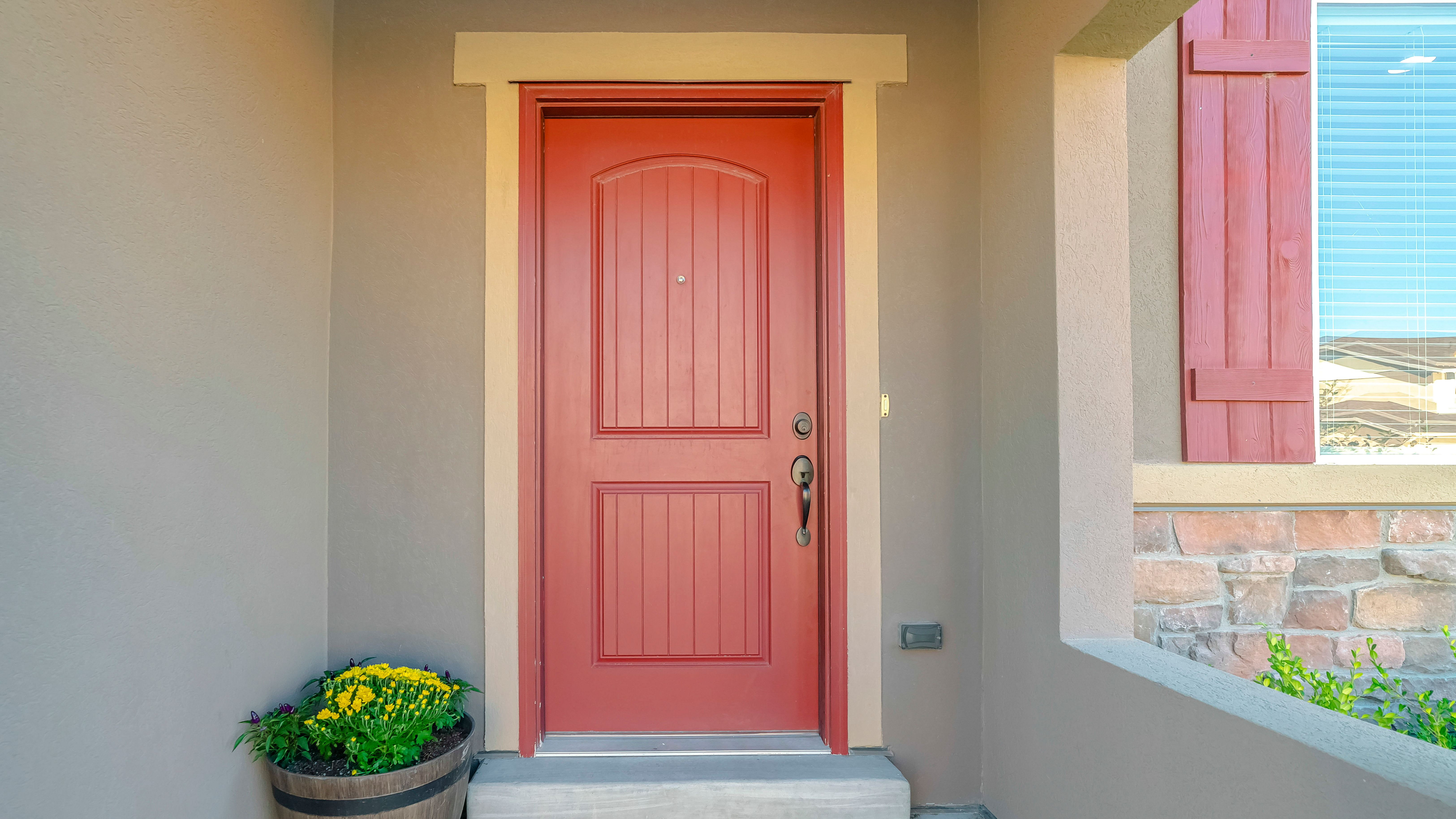 A vibrant coral-colored exterior door with a decorative window shutter beside it, set in a beige wall with a stone base.