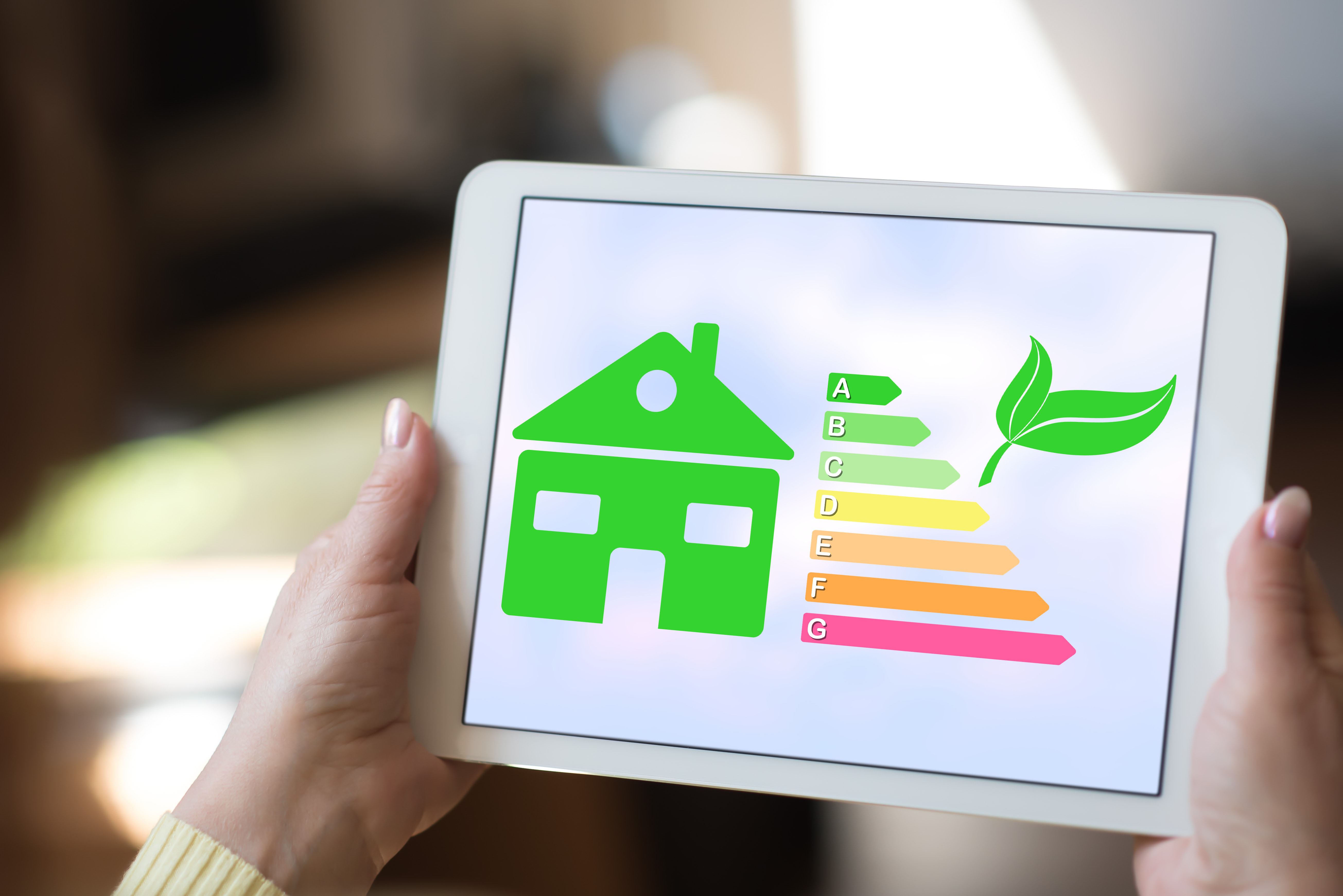 Hands holding a tablet displaying a green house icon with an energy efficiency rating chart.