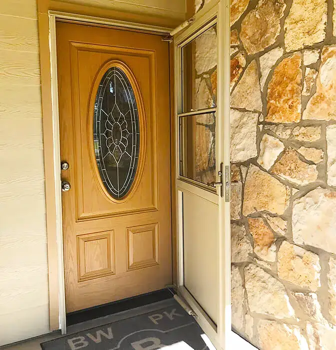 Elegant wooden front door with oval glass insert and white storm door, set against a rustic stone wall.