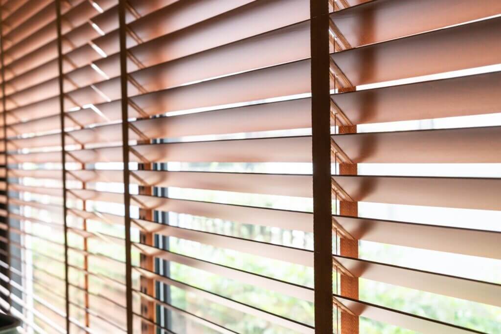 Sunlight filtering through brown wooden blinds revealing blurred greenery outside.