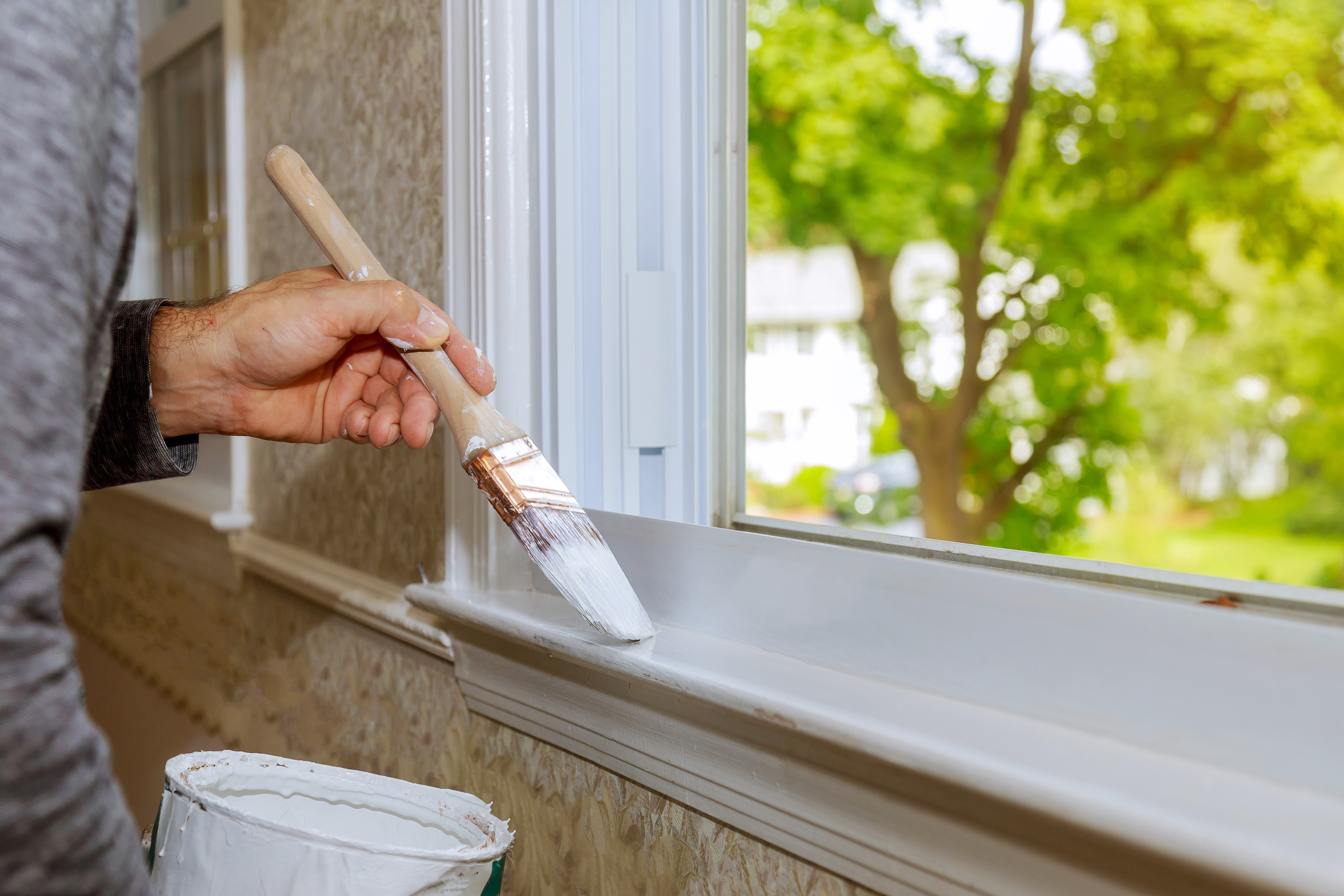 Hand applying white paint to a window frame, potentially leading to stuck windows.