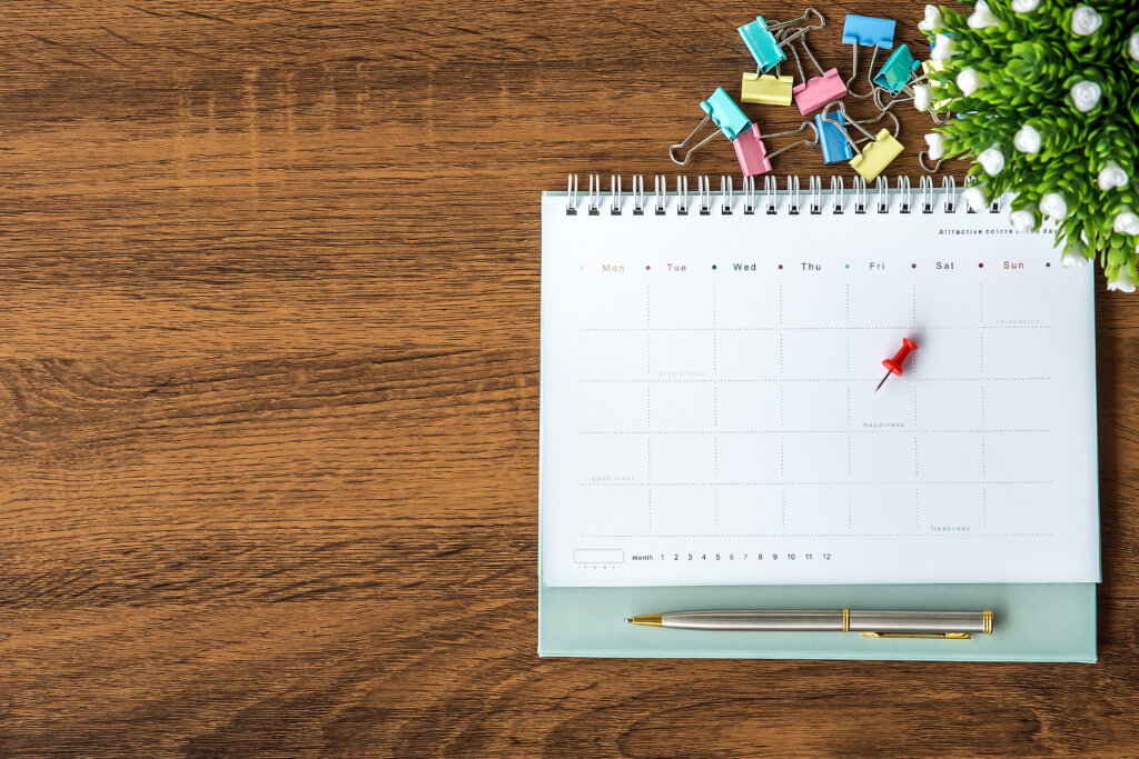 An open planner on a wooden desk with a red pushpin marking a date, surrounded by colorful binder clips and a pen.